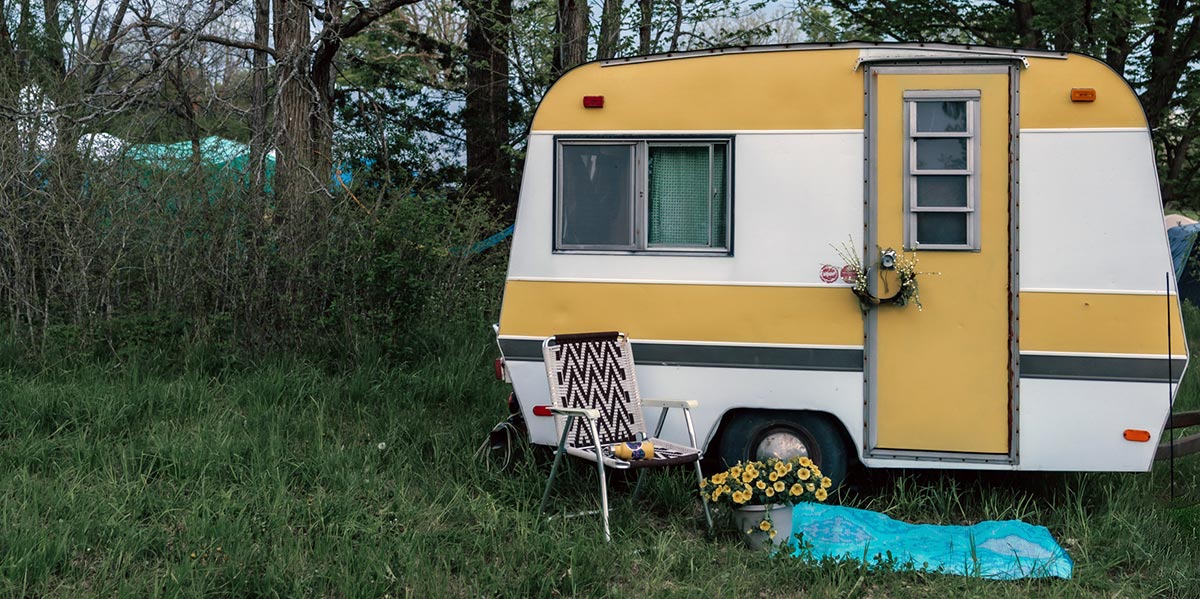 Image of a small vintage RV in a field of grass against a backdrop of trees.